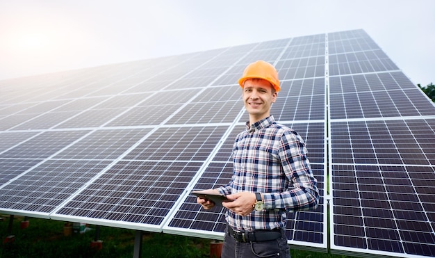 Glimlachende man in helm met tablet in handen staat in de buurt van zonnepanelen op groene plantage