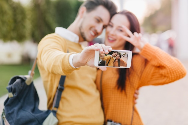 Glimlachende man met baard die witte smartphone vasthoudt en selfie maakt met schattig zwartharig meisje. Dromerige jonge vrouw in oranje gebreide kleding die zich voordeed op de achtergrond wazig natuur met vriendje.