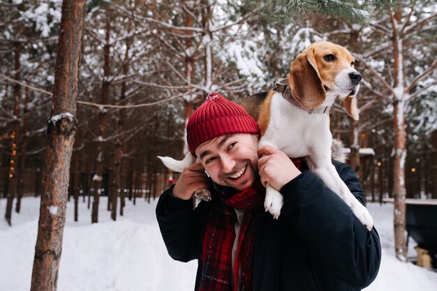 Glimlachende man speelt met een hond, ze ligt op zijn schouders, ze kijkt rustig in de verte. Buiten in een naaldboombos bij de winter.