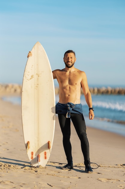 Glimlachende man surfer met naakte torso staande aan de kust met een surfplank