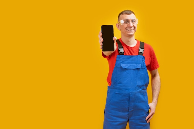 Glimlachende man werknemer in uniform en bril houdt slimme telefoon met leeg zwart scherm geïsoleerd op gele achtergrond Kopie ruimte mock up