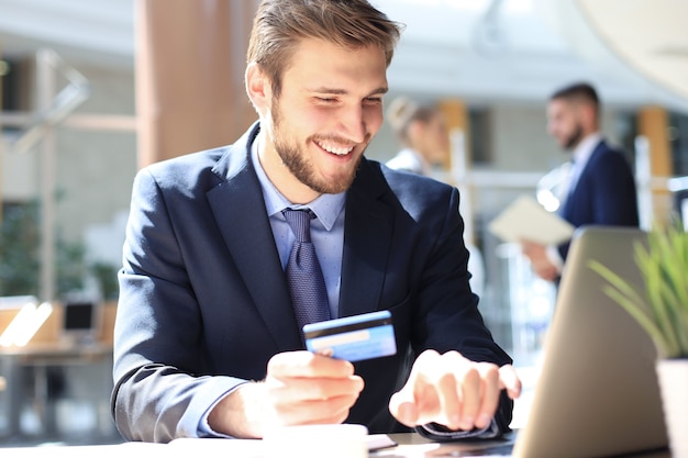 Glimlachende man zit op kantoor en betaalt met een creditcard met zijn laptop.