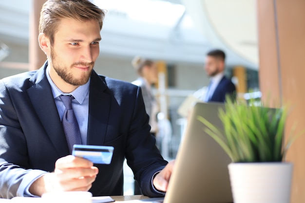 Glimlachende man zit op kantoor en betaalt met een creditcard met zijn laptop.