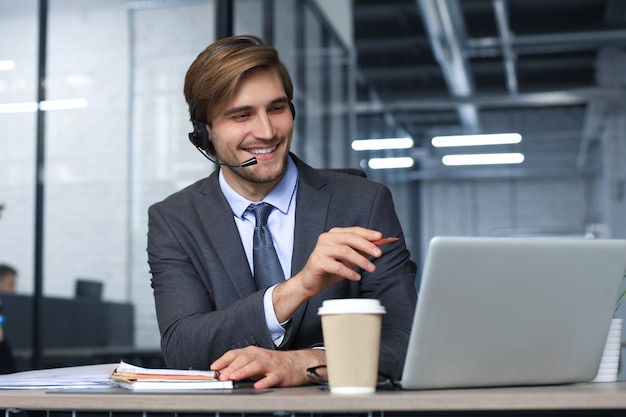 Glimlachende mannelijke callcenter-operator met koptelefoon zittend op een modern kantoor, online informatie raadplegend op een laptop, informatie opzoeken in een bestand om de klant te helpen.