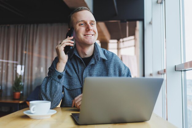 Glimlachende mannelijke ondernemer in vrijetijdskleding zittend aan tafel met laptop en pratend op smartphone in moderne werkruimte met groot raam overdag