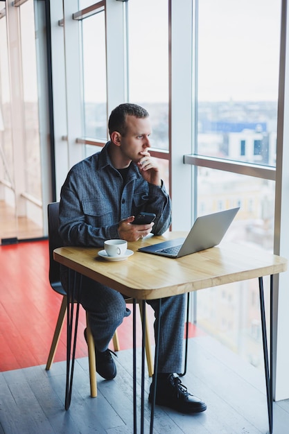 Glimlachende mannelijke ondernemer in vrijetijdskleding zittend aan tafel met laptop en pratend op smartphone in moderne werkruimte met groot raam overdag