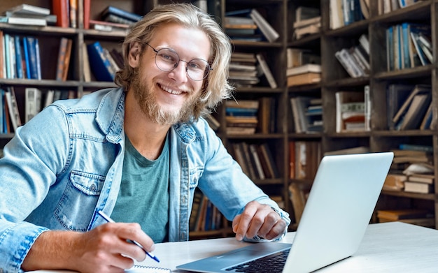 Glimlachende mannelijke student die laptopcomputer gebruikt en naar camera kijkt, leer gemakkelijk internetcursus, studeer online e-learning in app, schrijf aantekeningen, bereid je voor op proefexamen met apparaat, zit aan het bureau van de bibliotheek portret