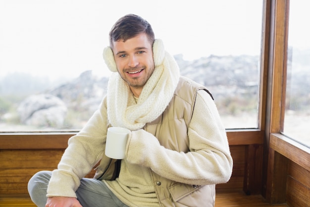 Glimlachende mens die oorbeschermer met koffiekop draagt ​​tegen venster