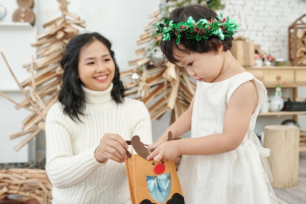 Glimlachende moeder die een gelukkig nieuwsgierig meisje bekijkt dat de zak opent met kerstcadeau