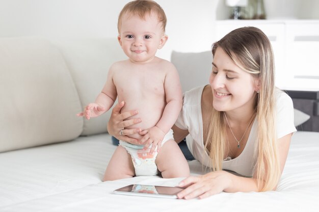 Glimlachende moeder en babyjongen die digitale tablet op bed gebruiken