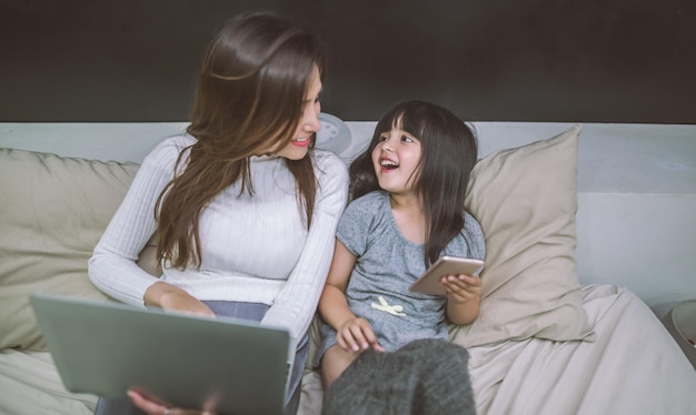 Glimlachende moeder en dochter gebruiken telefoon met laptop terwijl ze op bed zitten
