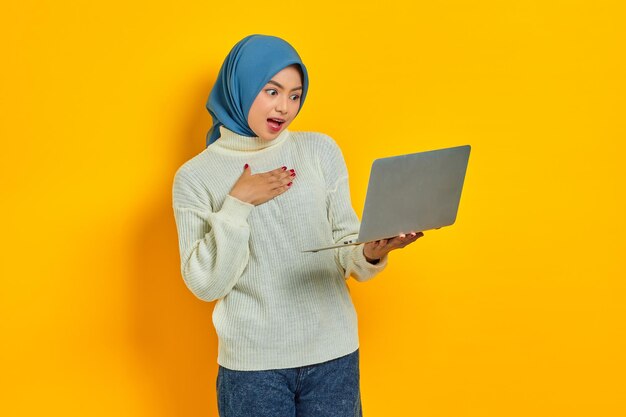 Glimlachende mooie Aziatische vrouw in witte trui met laptop met hand op borst geïsoleerd over gele achtergrond mensen religieuze levensstijl concept