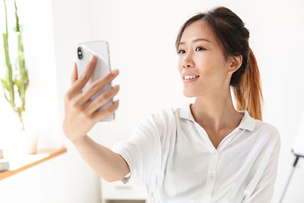 Glimlachende mooie jonge Aziatische zakenvrouw die een selfie neemt terwijl ze op kantoor staat