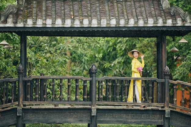 Glimlachende mooie jonge Vietnamese vrouw in gele jurk en hoed staande op brug met stelletje lo...