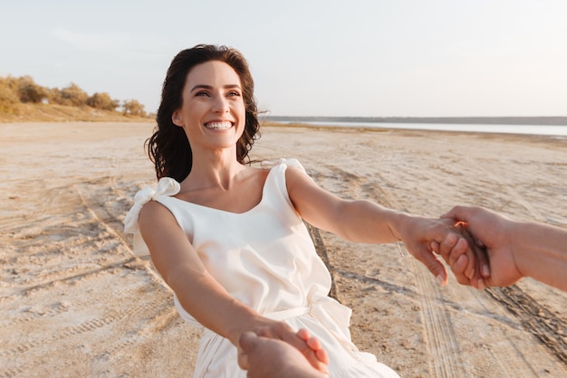 Glimlachende mooie jonge vrouw die een zomerjurk draagt en haar vriendjes uitgestrekte hand vasthoudt tijdens het wandelen op het strand