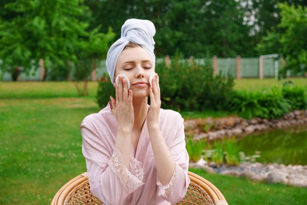 Glimlachende mooie jonge vrouw met een handdoek op het hoofd houdt een wattenschijfje vast om het gezicht te reinigen