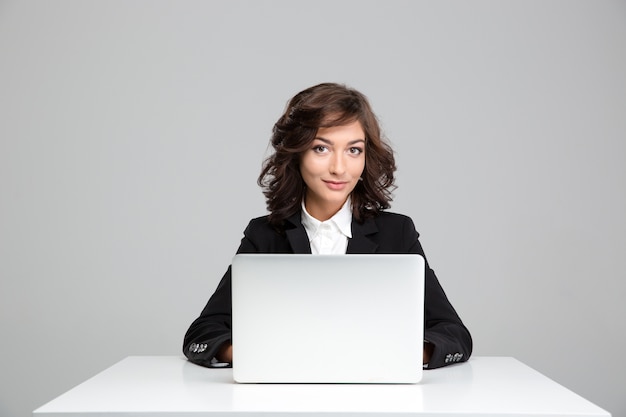 Glimlachende mooie krullende jonge vrouw in zwarte jas zitten en laptop gebruiken