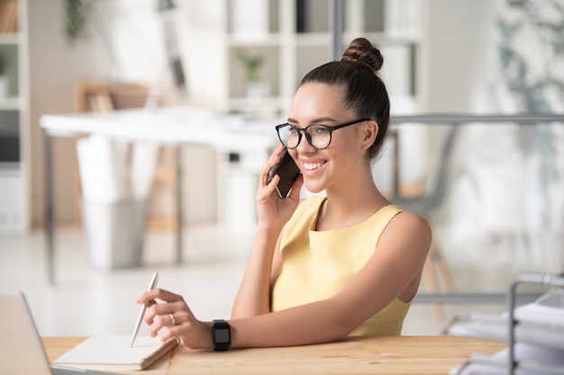 Glimlachende mooie onderneemster met haarbroodje die aan bureau zitten en verder project telefonisch bespreken