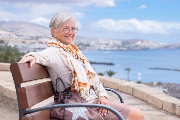 Glimlachende mooie oudere vrouw met bril die buiten op een bankje zit en naar zee kijkt en geniet van vrijheid en vakantie Actieve oudere dame die ontspant op een zonnige dag