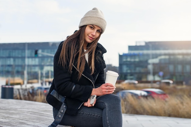 Glimlachende mooie vrouw met smartphone en koffie om buiten te gaan staan en modern te spreken