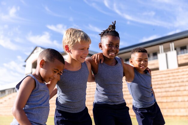 Foto glimlachende multiraciale elementaire jongens die zich met arm rond tegen schoolgebouw op zonnige dag