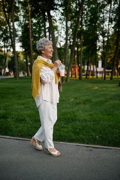 Glimlachende oma houdt melkcocktail vast in het zomerpretpark, terug naar de kindertijd. leeftijd mensen levensstijl. grappige grootmoeder die buiten plezier heeft, oude vrouwelijke persoon op de natuur