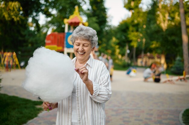 Glimlachende oma houdt suikerspin in zomerpretpark. Leeftijd mensen levensstijl. Grappige grootmoeder die buiten plezier heeft, oude vrouwelijke persoon op de natuur