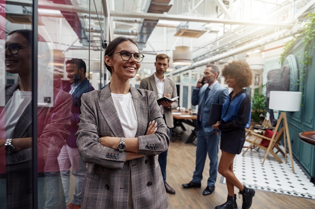 Glimlachende onderneemster in glazen die zich in modern bureau op collega'sachtergrond bevinden