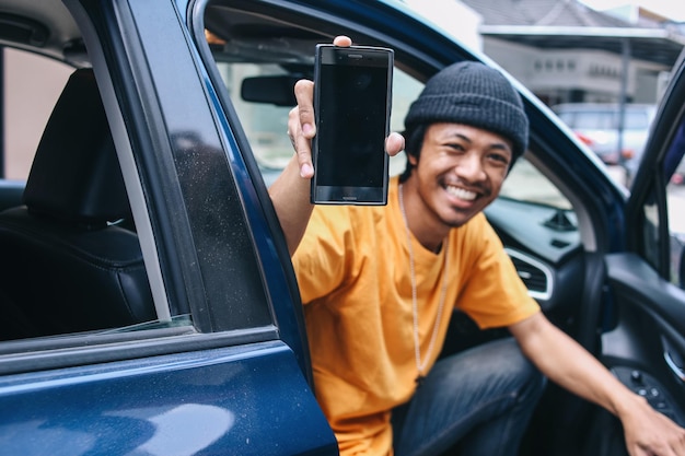 Glimlachende online taxichauffeur die een leeg telefoonscherm toont voor mockup terwijl hij in de auto zit