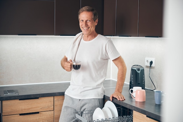 Glimlachende ontspannen volwassen man die in de keuken staat en geniet van zijn aromakoffie