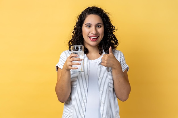 Glimlachende optimistische vrouw die zich met glas water bevindt dat duim als drank toont