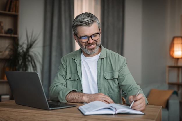 Glimlachende oudere Europese man met baard die met laptop werkt, bekijk videoles of webinar in de woonkamer