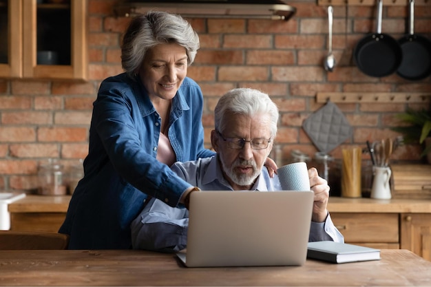 Glimlachende oudere rijpe grijze vrouw die fijner op laptopscherm wijst en vakantiereis kiest met ontspannen senior echtgenoot online Gelukkig familiepaar van middelbare leeftijd dat moderne tech-gadget samen thuis gebruikt