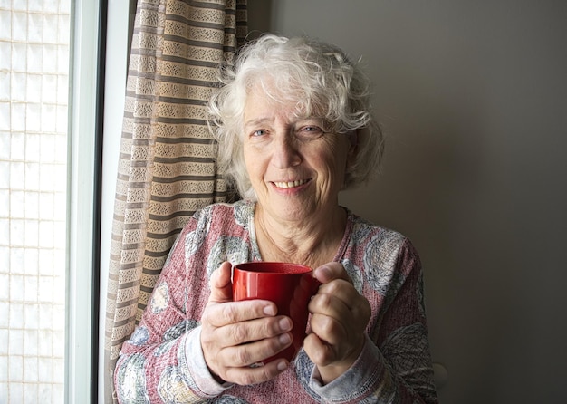 Foto glimlachende oudere vrouw die met een kop koffie in haar handen staat en naar de camera kijkt