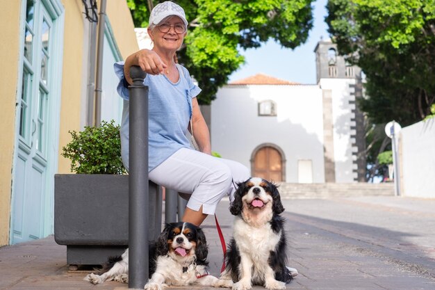 Glimlachende oudere vrouw met hoed die op straat zit met haar twee cavalier koning Charles spaniel honden