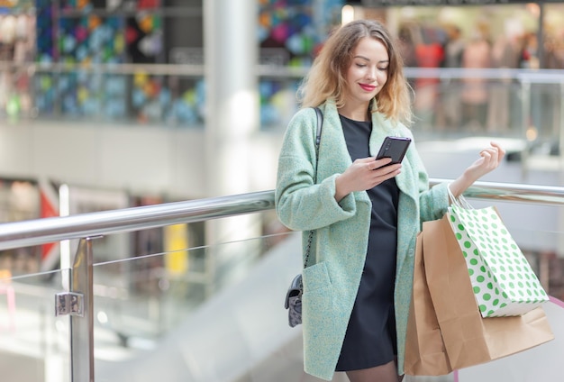 Glimlachende, over het algemeen schattige blanke vrouw met bretels en krullend haar gebruikt telefoon met boodschappentassen in het winkelcentrum