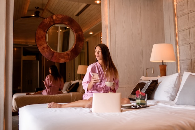 Glimlachende perfecte vrouw in roze badjas met kopje thee / koffie en laptop zitbed in de gezellige hotelkamer.