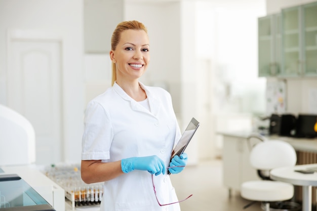 Glimlachende positieve aantrekkelijke vrouwelijke labassistent met rubberen handschoenen in steriele uniforme bril en tablet te houden terwijl je in het laboratorium staat.