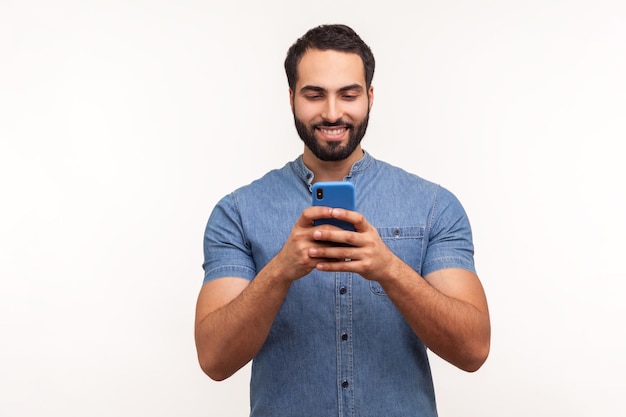 Glimlachende positieve bebaarde man in blauw shirt kijkend naar smartphone-display, surfen op internet, wedden, online winkelen. Indoor studio-opname geïsoleerd op een witte achtergrond