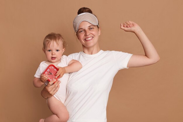 Glimlachende positieve krachtige vrouw die wit t-shirt en blinddoek draagt die babydochter met rode wekker in kleine handen houdt die met goed humeur wakker wordt moeder opgeheven arm die laat zien hoe sterk ze is