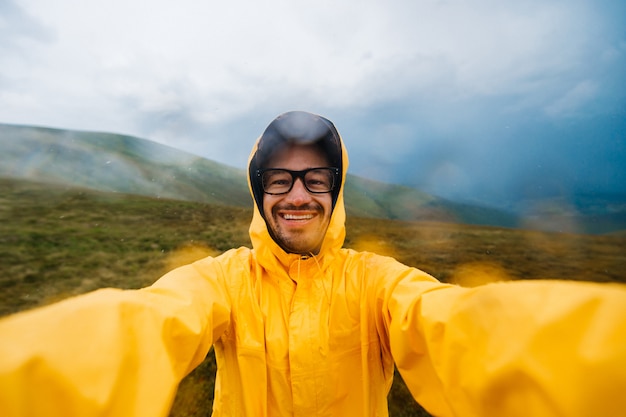 Glimlachende reizigersmens in gele regenjas die selfie in regenachtige bergen nemen.