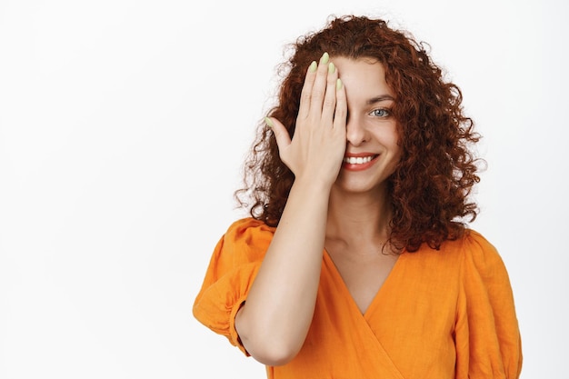 Glimlachende roodharige krullende vrouw, bedek een kant van het gezicht en kijk gelukkig, glimlach naar de camera, staande in een oranje jurk tegen een witte achtergrond. Ruimte kopiëren