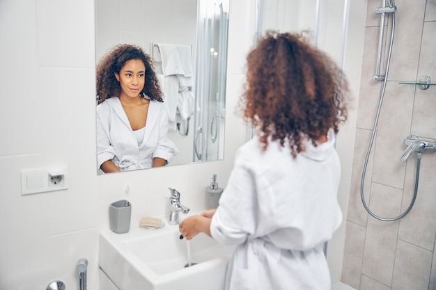 Foto glimlachende schattige jonge vrouw die voor de kraan staat met stromend water