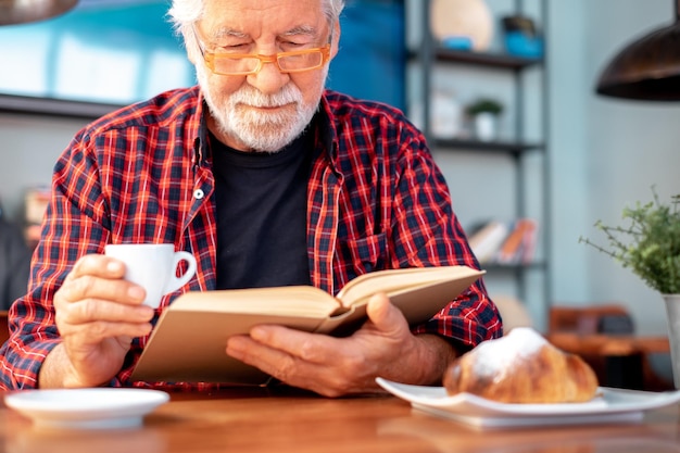 Glimlachende senior bebaarde man zittend aan cafétafel met een boek genietend van leesplezier en pauze