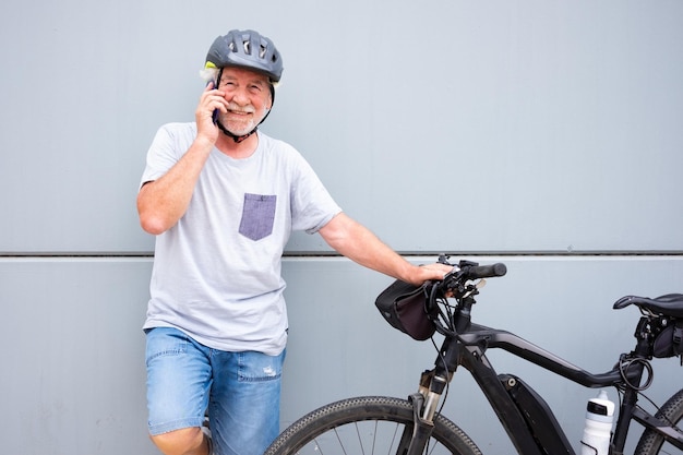 Glimlachende senior fietser man met helm met behulp van telefoon dicht bij zijn elektrische fiets