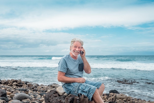 Glimlachende senior man genieten van ontspanning en vrijheid op het strand zitten met behulp van smartphone