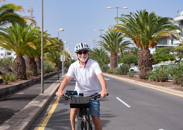 Glimlachende senior man met helm genieten van gezonde levensstijl lopen met zijn elektrische fiets in een zonnige stad straat palmbomen op achtergrond duurzaam mobiliteitsconcept