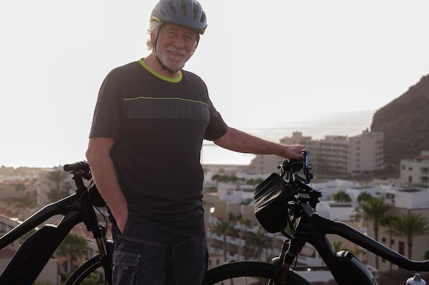 Glimlachende senior sportieve man met helm staande op heuvel met elektrische fiets gezonde activiteit voor gepensioneerde man genieten van vakantie