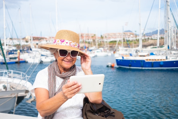 Glimlachende senior vrouw die buiten in de haven zit, neemt een selfie met digitale tablet