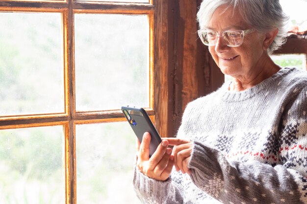 Glimlachende senior vrouw in wintertrui met behulp van mobiele telefoon bij het raam in een rustiek chalet